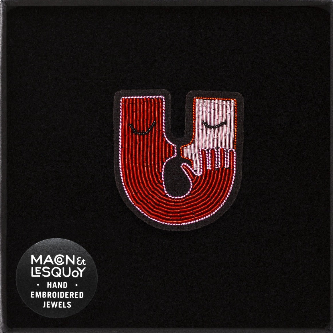 Broche brodée à la main en cannetille de la marque Macon et Lesquoy. Cette broche est en forme de U représentant deux visages en face à face de couleur rouge et rose qui se font un bisou