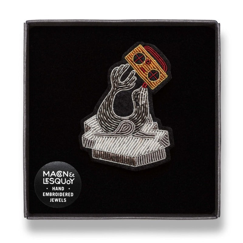 Broche brodée à la main en fil de cannetille de la marque Macon & Lesquoy. Collection Froid Automne-Hiver 2022. Broche musique de loutre. Coloris : argenté et nuances de doré et marron. Dimensions : 4,1 x 5,8 cm. broche dans son écrin de velours noir.