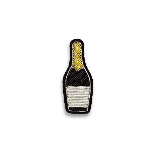 Broche brodée à la main de la marque Macon et Lesquoy. Cette broche de couleur blanche, noire et dorée représente une bouteille de champagne. 