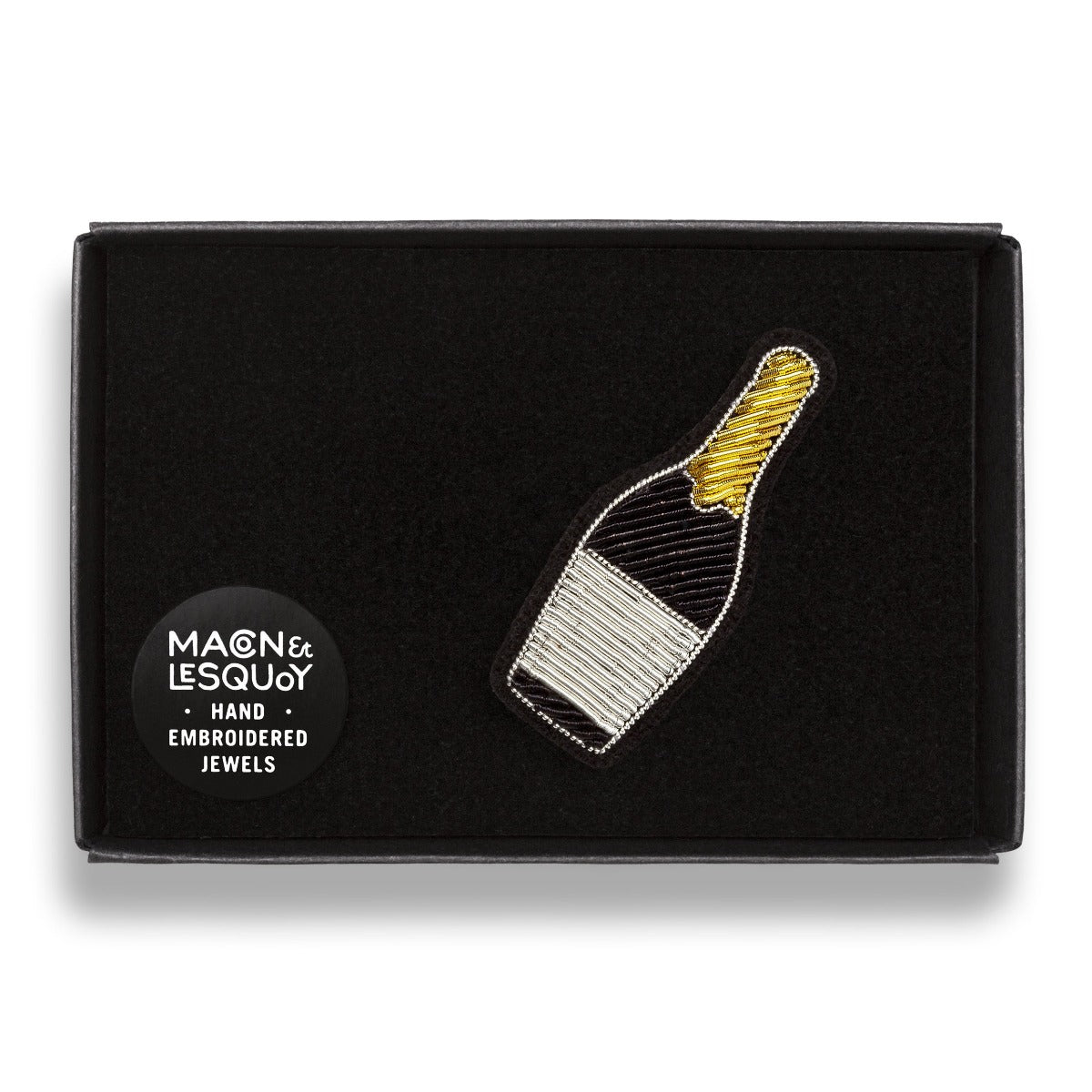 Broche brodée à la main de la marque Macon et Lesquoy. Cette broche de couleur blanche, noire et dorée représente une bouteille de champagne.