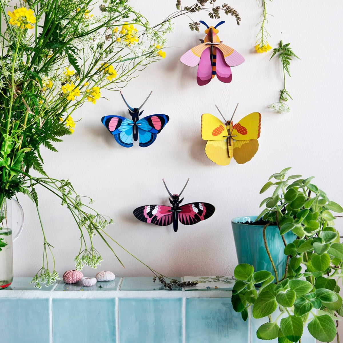 Un univers de papillons en carton recyclé de la marque studio roof, un bleu, un rose, un jaune et un fushia accrochés au mur entouré de fleurs coupées et de plantes.